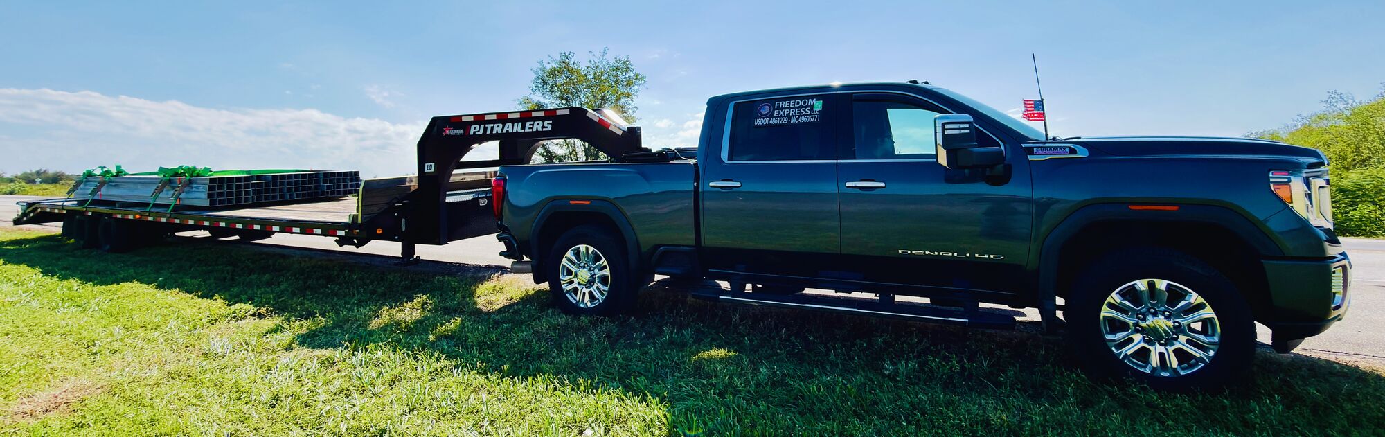 A dark GMC Sierra Denali HD lettered Freedom Express LLC, USDOT 4861229, MC 49605771, hitched to a PJ Trailers gooseneck flatbed loaded with strapped-down steel tubing